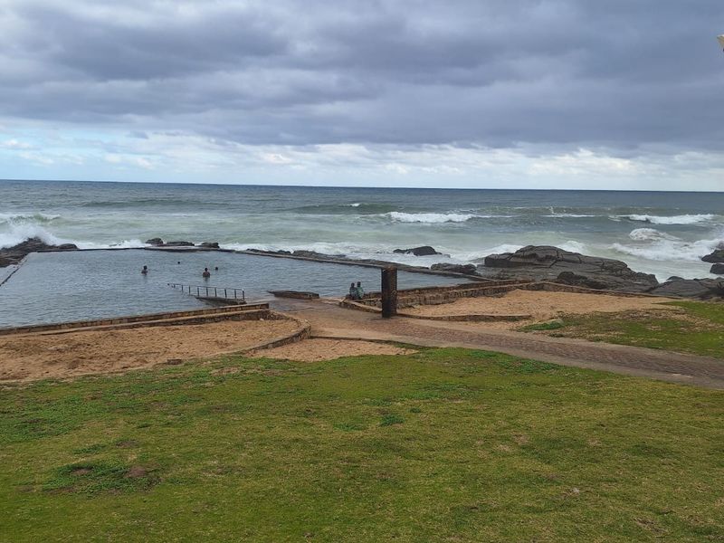 A coastal outdoor pool area featuring a natural rock-edge swimming pool overlooking the ocean with adjacent paved pathways and lawn.