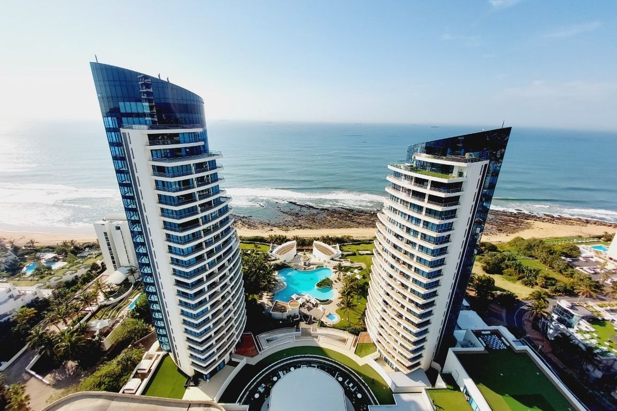 Aerial view of a luxury beachfront apartment complex featuring twin high-rise towers, a central swimming pool, and landscaped grounds overlooking the ocean.