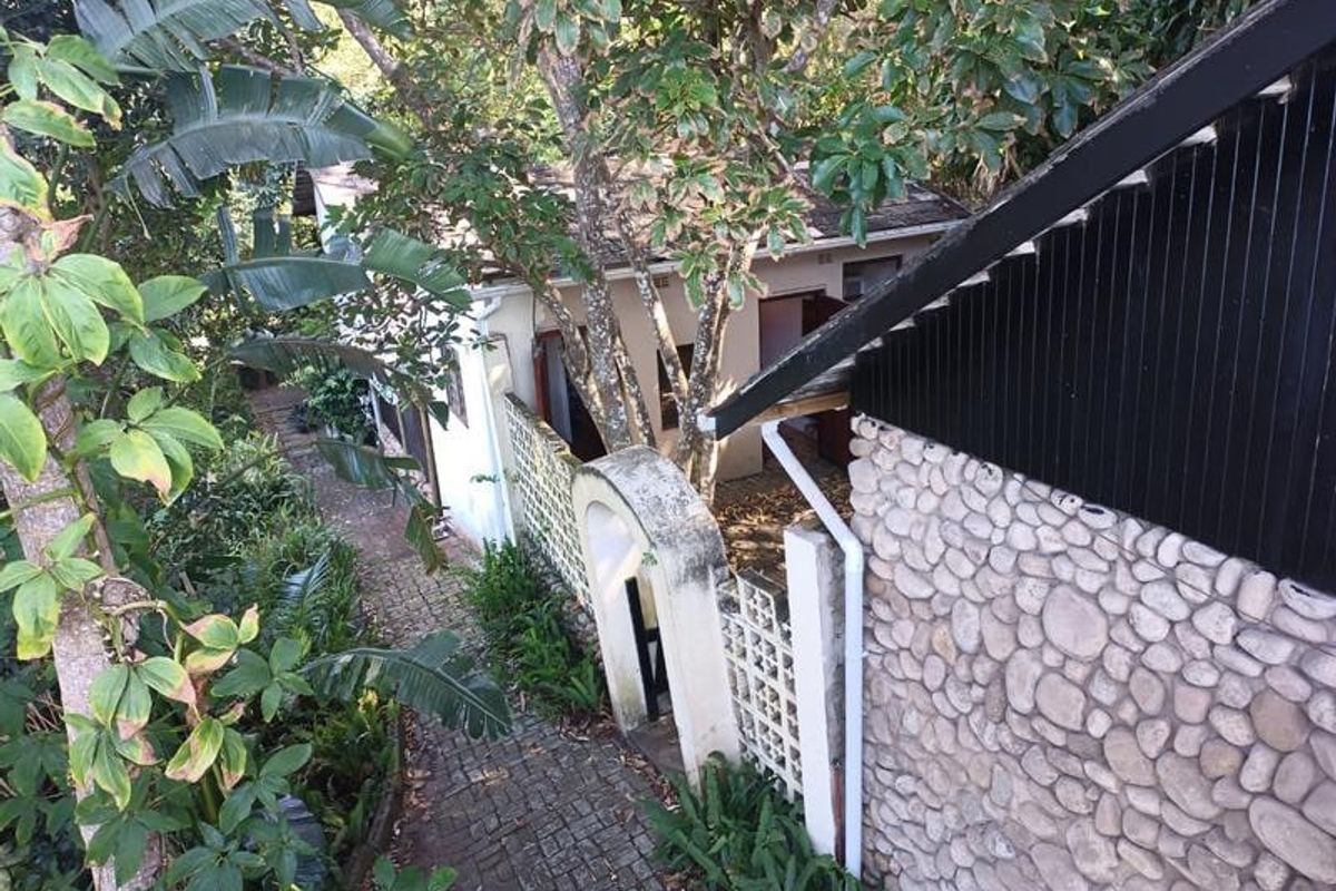 A rear exterior view showing a stone-clad wall, a paved walkway leading to an arched gate, and mature garden landscaping.