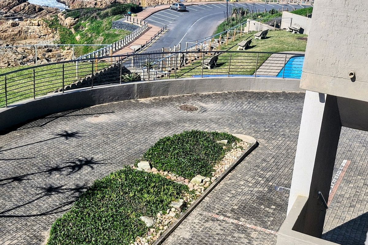 Elevated view of the apartment complex exterior, featuring a paved driveway, landscaped garden beds, metal balustrades, and a communal swimming pool area.