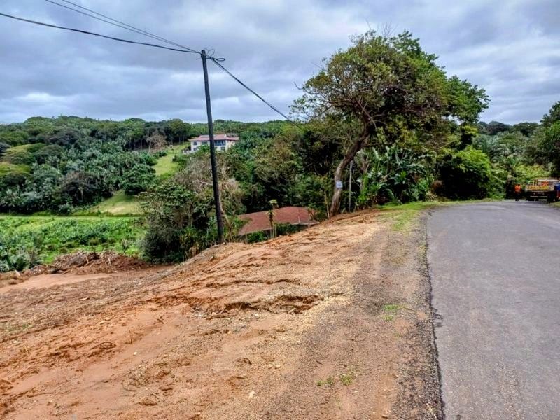 A view of the vacant residential land plot adjacent to an asphalt road, featuring sloping terrain and surrounding natural vegetation.