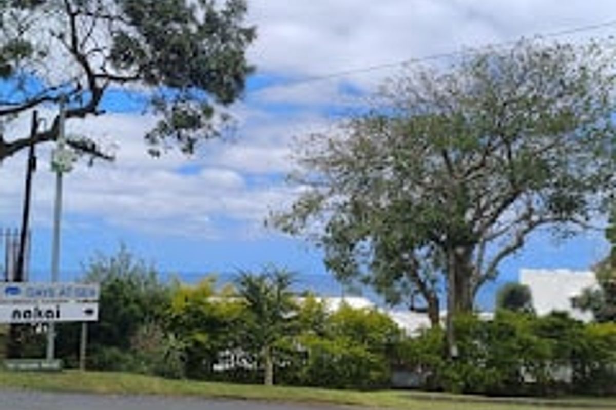 A street-level view of the property frontage featuring mature trees and local signage, with a glimpse of the surrounding landscape.