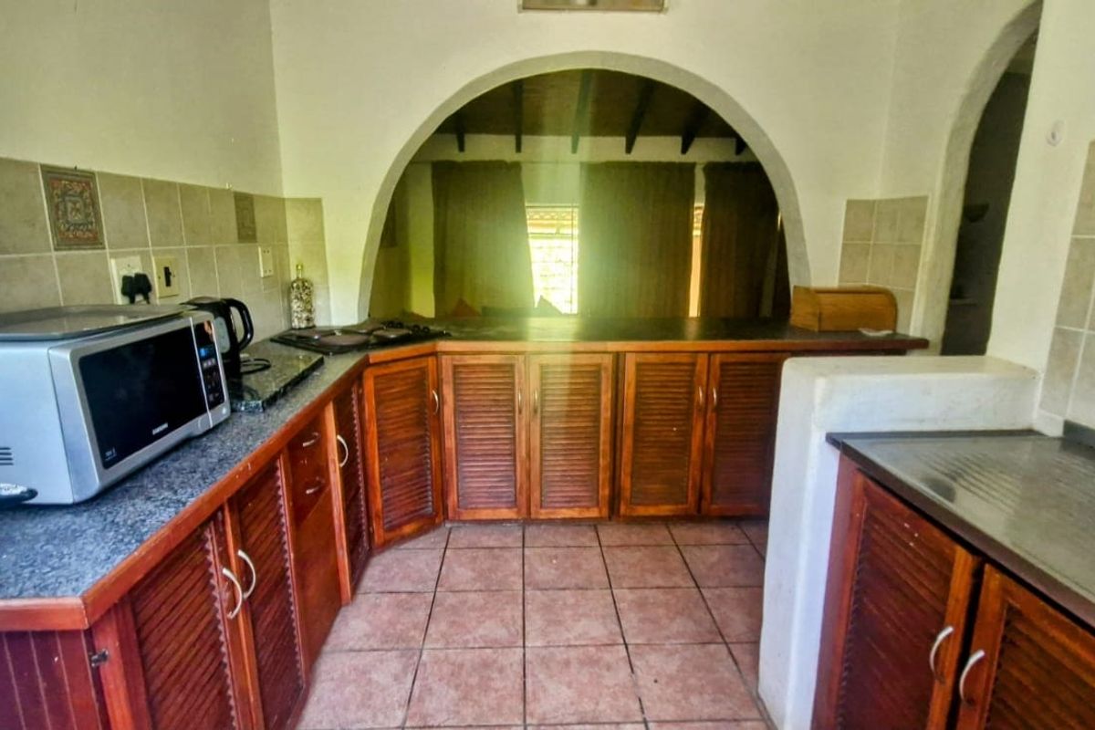 A dated kitchen featuring louvered timber cabinetry, tiled splashbacks, and ceramic flooring, connected to an adjacent living space via a structural archway.