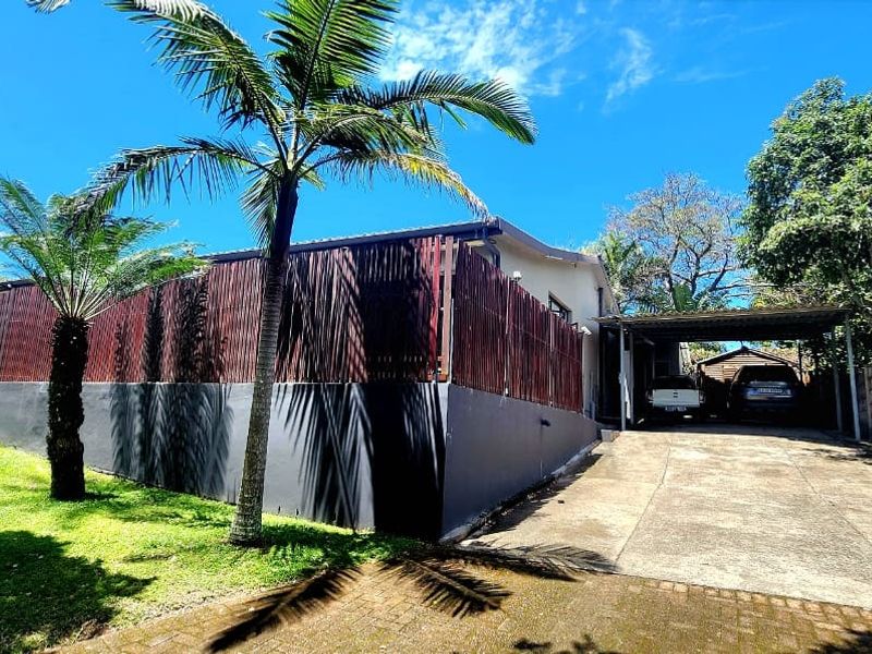 Front exterior view featuring a concrete driveway, covered carport, timber slat fencing, and a landscaped garden area with mature palm trees.