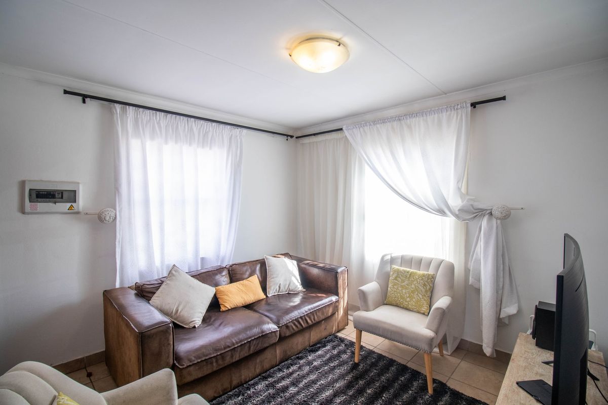 A cozy living room featuring tiled flooring, dual windows with curtain rods, and a wall-mounted electrical distribution board.