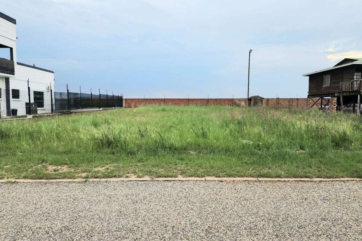 A vacant residential plot featuring a level grass surface, enclosed by a brick boundary wall with electric fencing, situated between existing properties.