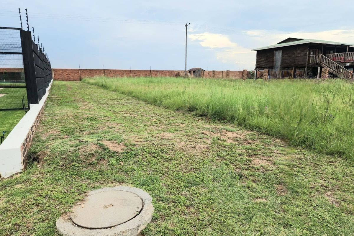 A view of a vacant residential plot featuring a brick boundary wall, electric fencing, and an existing structure in the background.