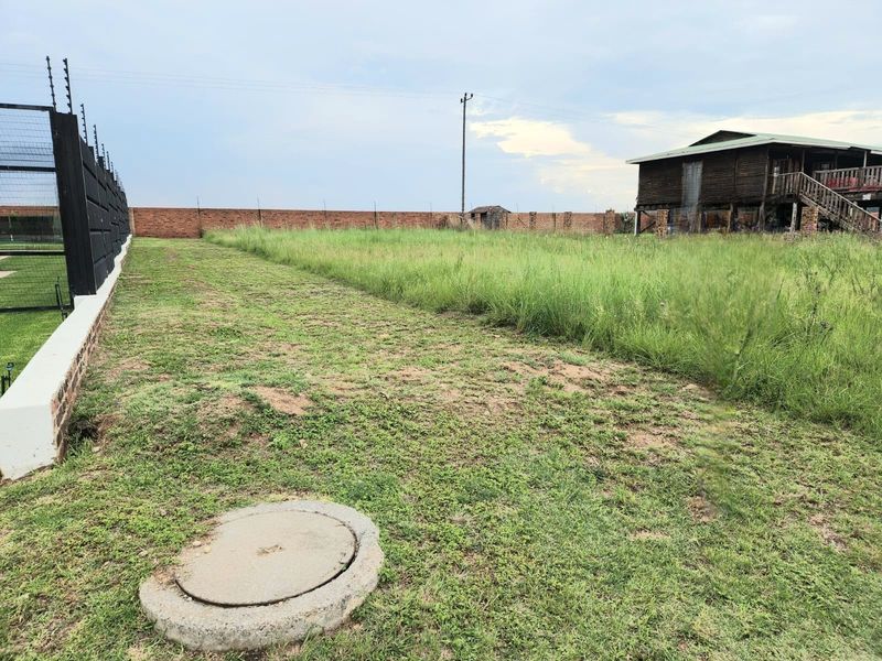 A view of a vacant residential plot featuring a brick boundary wall, electric fencing, and an existing structure in the background.