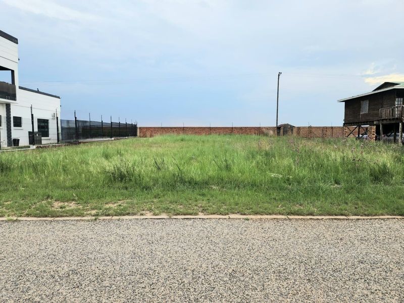 A vacant residential plot featuring a level grass surface, enclosed by a brick boundary wall with electric fencing, situated between existing properties.
