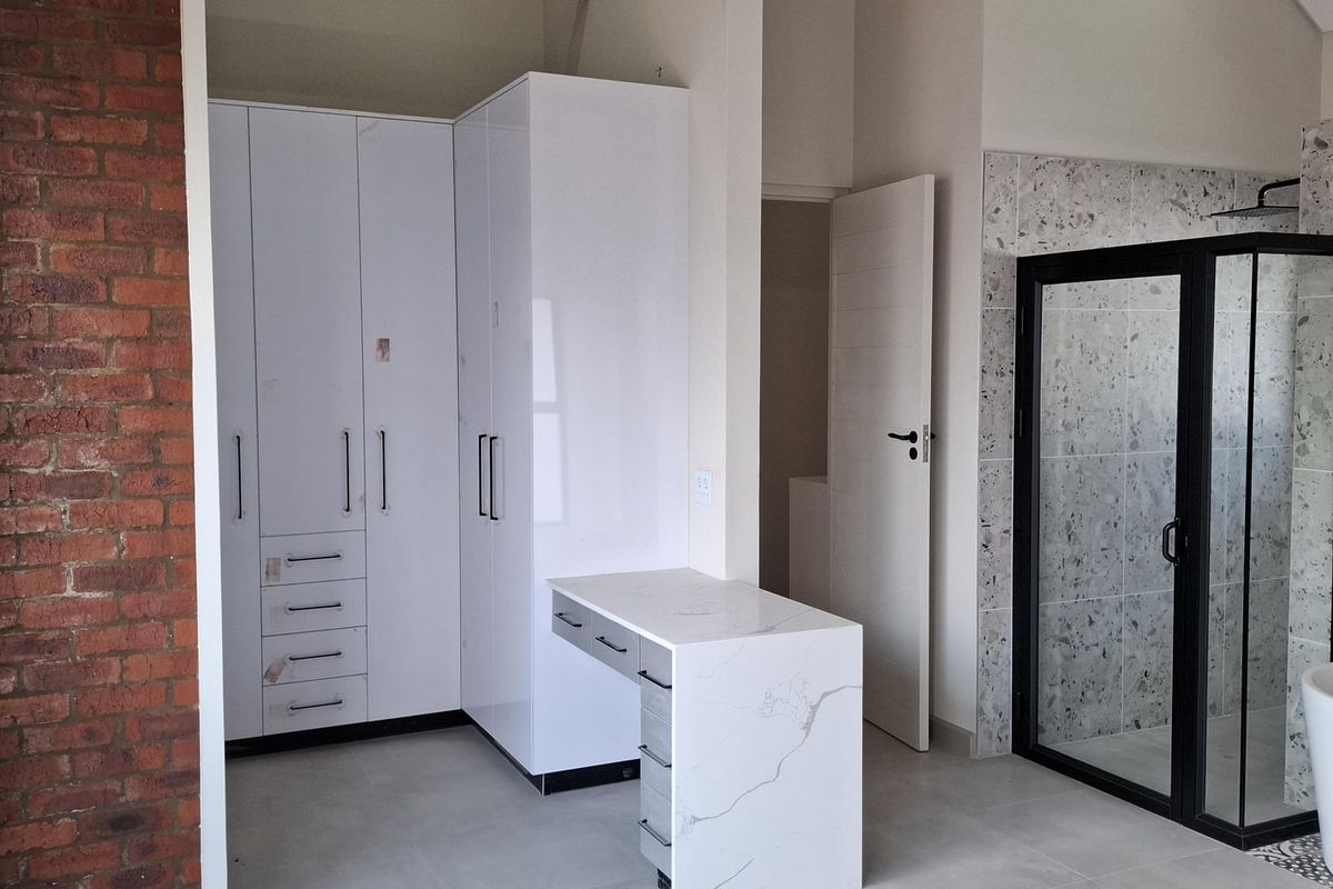 Spacious bedroom featuring exposed timber trusses, a brick feature wall, modern built-in cabinetry with an integrated vanity, and an en-suite walk-in shower.