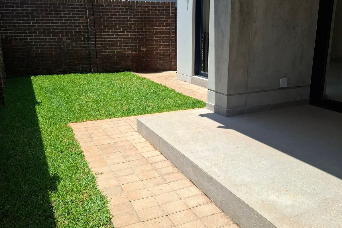 A private rear garden featuring a neat lawn, paved walkway, and a covered concrete patio area enclosed by a brick boundary wall.