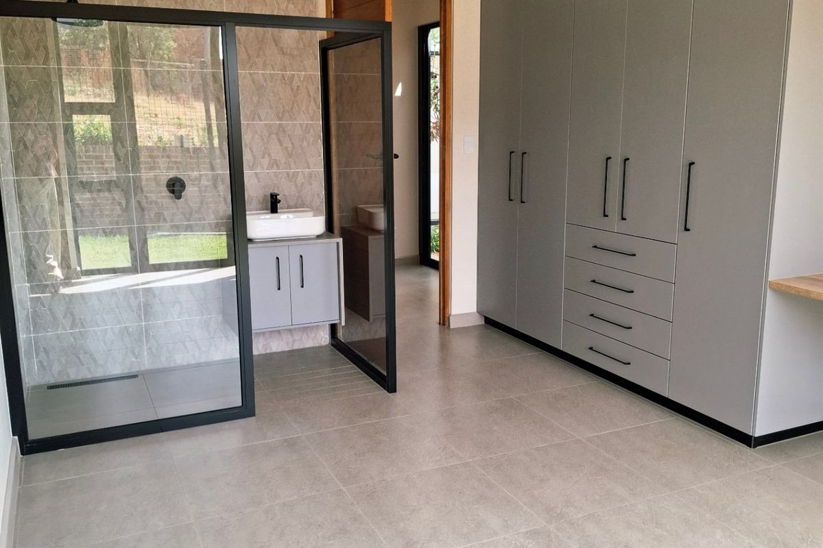 A modern bedroom featuring extensive built-in grey cabinetry and an open-plan en-suite bathroom with a glass-enclosed shower and vanity.
