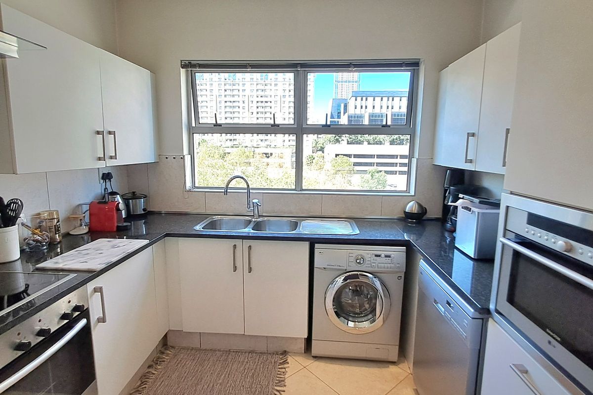 A functional kitchen featuring white cabinetry, dark countertops, built-in oven, hob, and integrated appliances with a window overlooking the city.