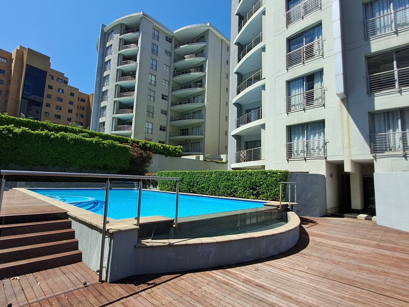 Outdoor pool area featuring timber decking, a glass balustrade, and manicured hedges, set against the backdrop of a modern multi-story apartment complex.