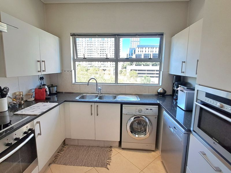A functional kitchen featuring white cabinetry, dark countertops, built-in oven, hob, and integrated appliances with a window overlooking the city.