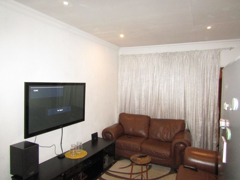 A compact living room featuring recessed downlighting and coving, with a wall-mounted television and full-length curtains.