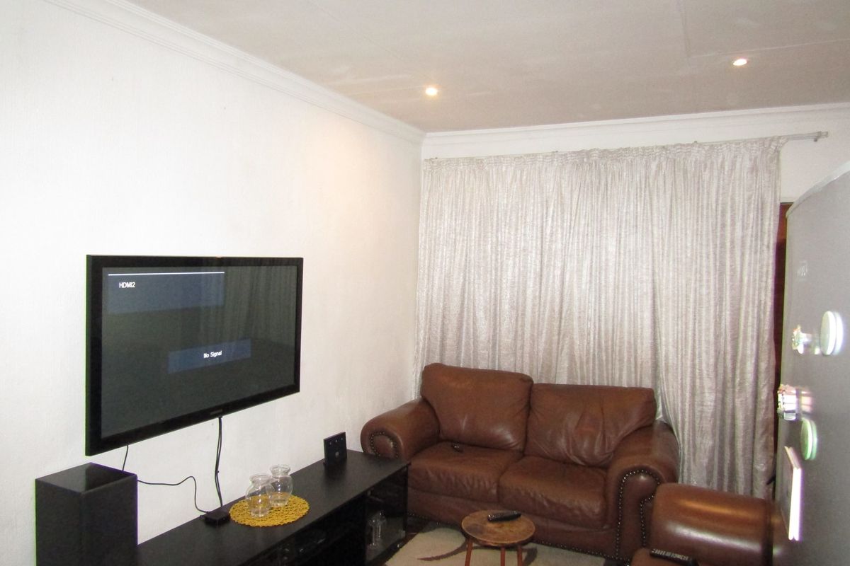 A compact living room featuring recessed downlighting and coving, with a wall-mounted television and full-length curtains.