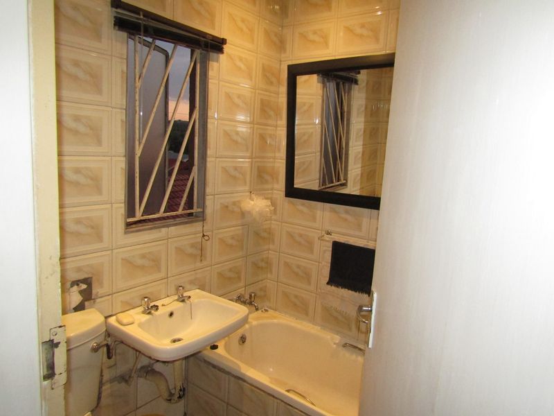 A compact bathroom featuring a bathtub, pedestal sink, and toilet with dated wall tiling and security bars on the window.