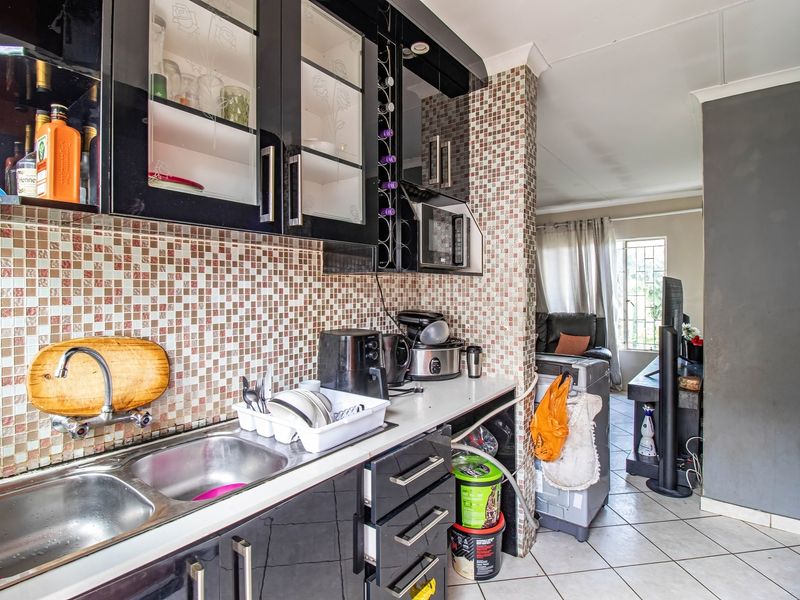 A compact kitchen featuring black cabinetry, mosaic tile backsplash, and a double bowl stainless steel sink, opening into an adjacent living area.
