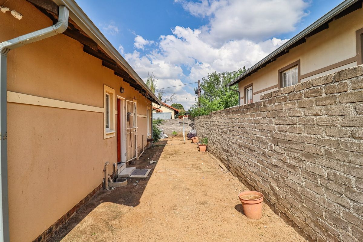 A narrow rear exterior space featuring a brick boundary wall, unpaved ground, and access to the side of the townhouse structure.