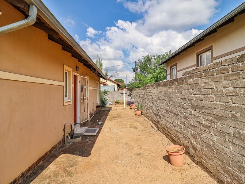 A narrow rear exterior space featuring a brick boundary wall, unpaved ground, and access to the side of the townhouse structure.