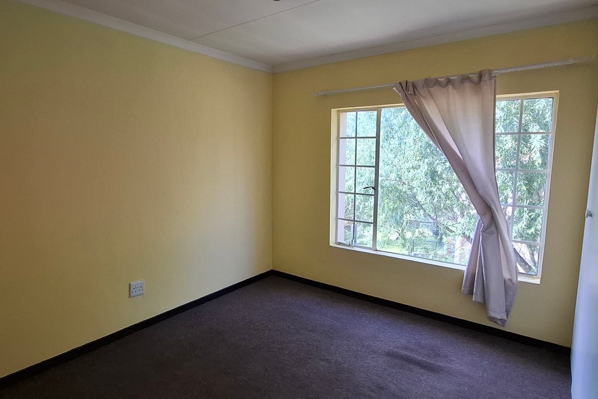 A bright, empty bedroom featuring carpeted flooring, a built-in wardrobe, and a window with security bars overlooking greenery.