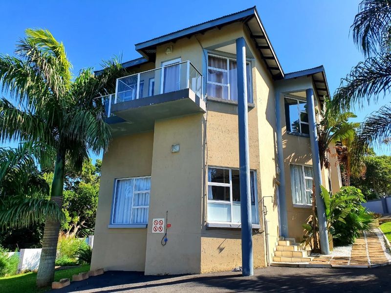 A two-story residential exterior featuring a balcony with glass balustrades, tiled entrance steps, and a paved driveway surrounded by established garden greenery.