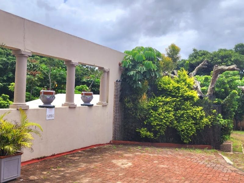 An outdoor rear area featuring a decorative colonnade wall, brick paved surface, and established garden landscaping.