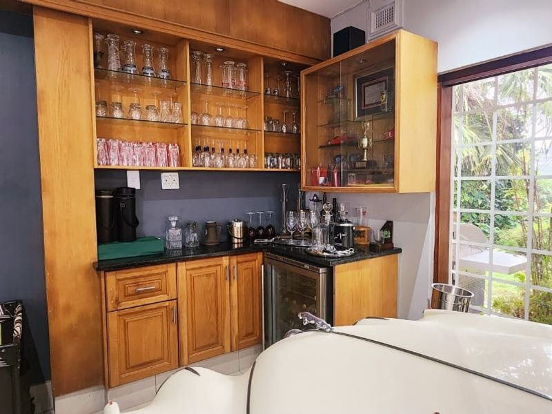 A dedicated home bar area featuring custom wooden cabinetry, glass display shelving, granite countertops, and an integrated under-counter beverage fridge with garden views.