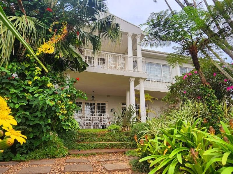 Rear view of a two-story house featuring a covered patio, upper balcony with columns, and a lush, well-established garden with a stone pathway.