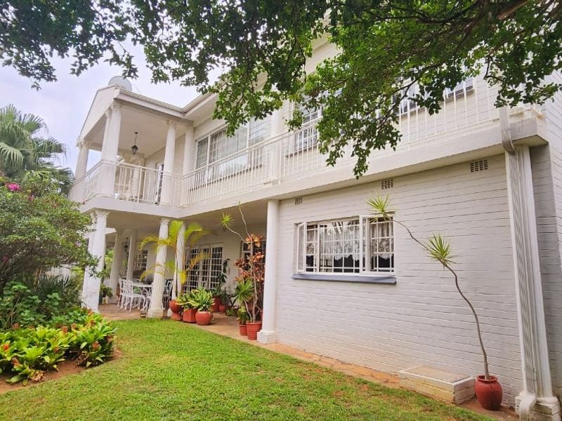 Rear view of a two-story painted brick house featuring a covered ground floor patio, an upper-level balcony, and established garden surroundings.