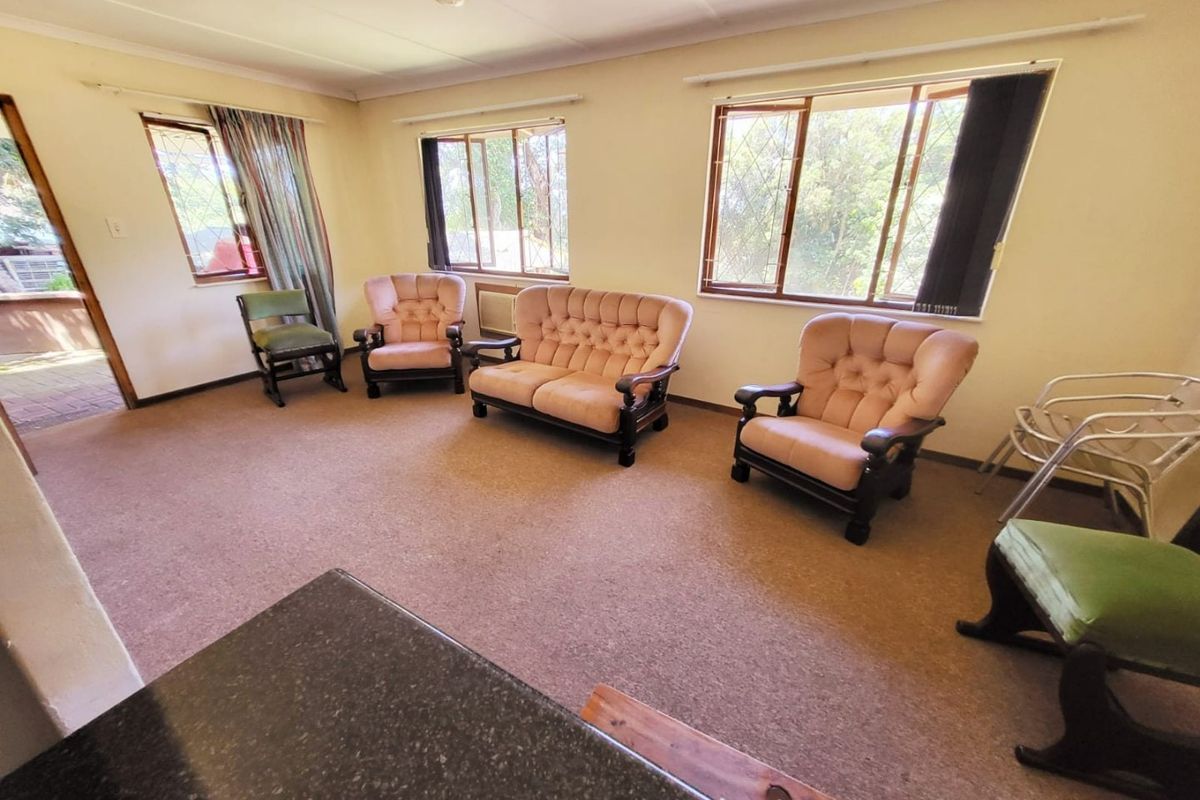 A spacious living room featuring carpeted flooring, coved ceilings, and large windows with security bars, offering a dated but functional layout.