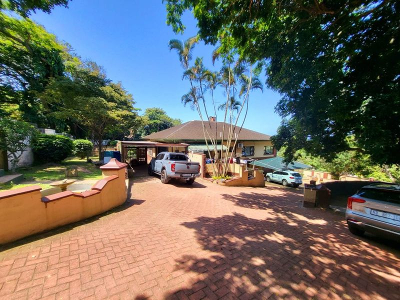 Front exterior view of the property featuring a spacious brick-paved driveway, carport, and established garden with mature trees.