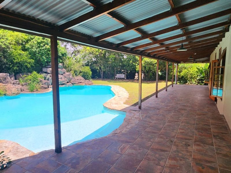 Spacious covered patio overlooking a landscaped swimming pool area with natural rock features and lush garden surroundings.