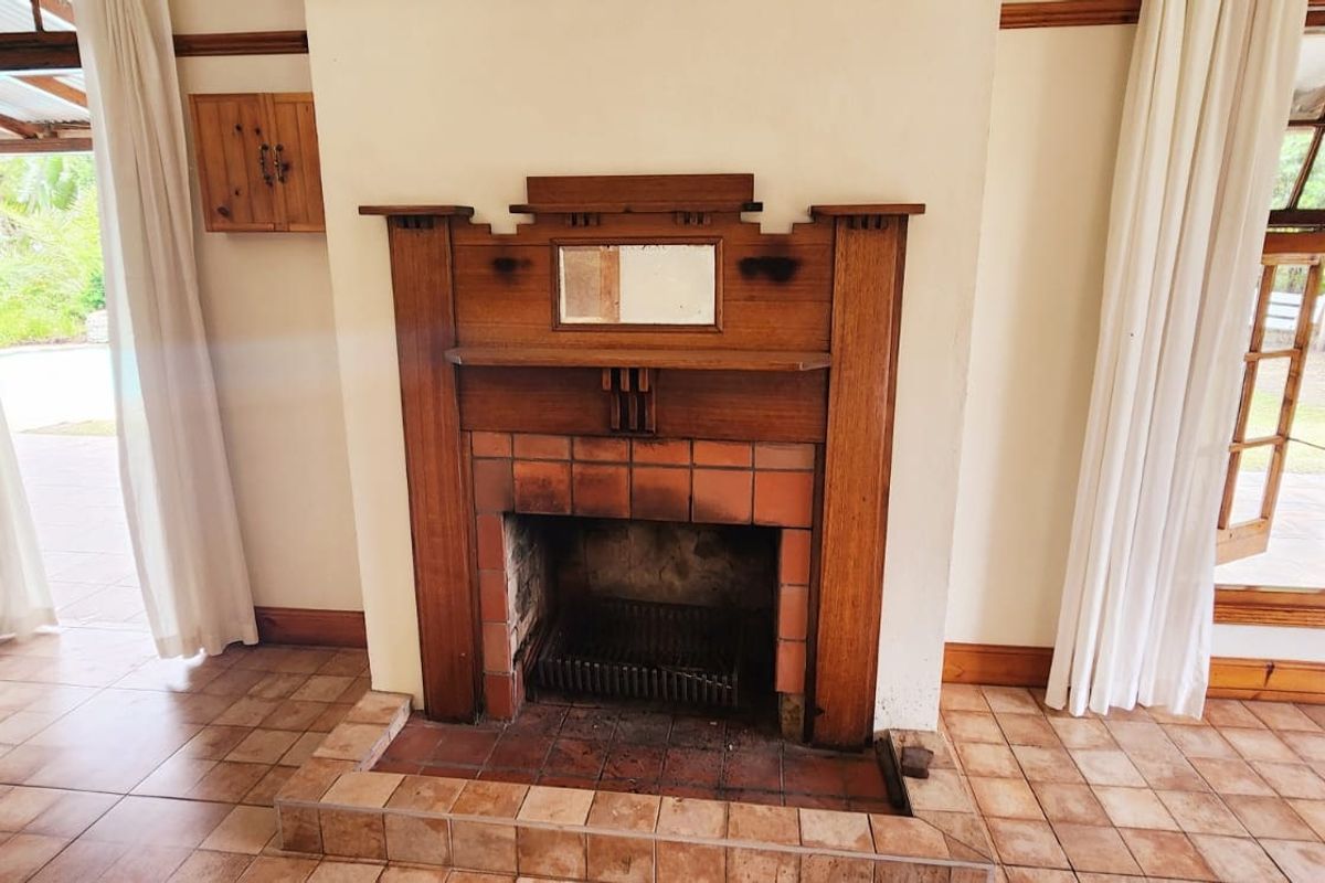 A living room featuring a traditional wooden fireplace mantel, tiled flooring, and large timber-framed windows and doors providing access to the outdoor area.