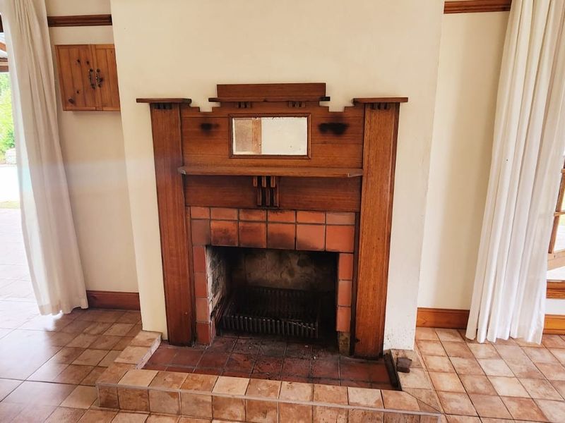 A living room featuring a traditional wooden fireplace mantel, tiled flooring, and large timber-framed windows and doors providing access to the outdoor area.