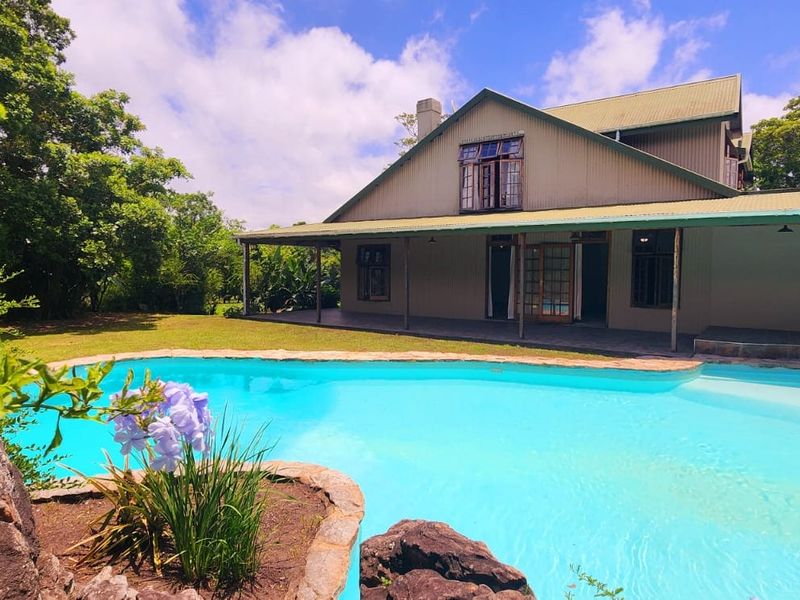 A well-maintained farmhouse featuring a large swimming pool, stone-edged garden beds, and a spacious covered patio area overlooking the lush property grounds.