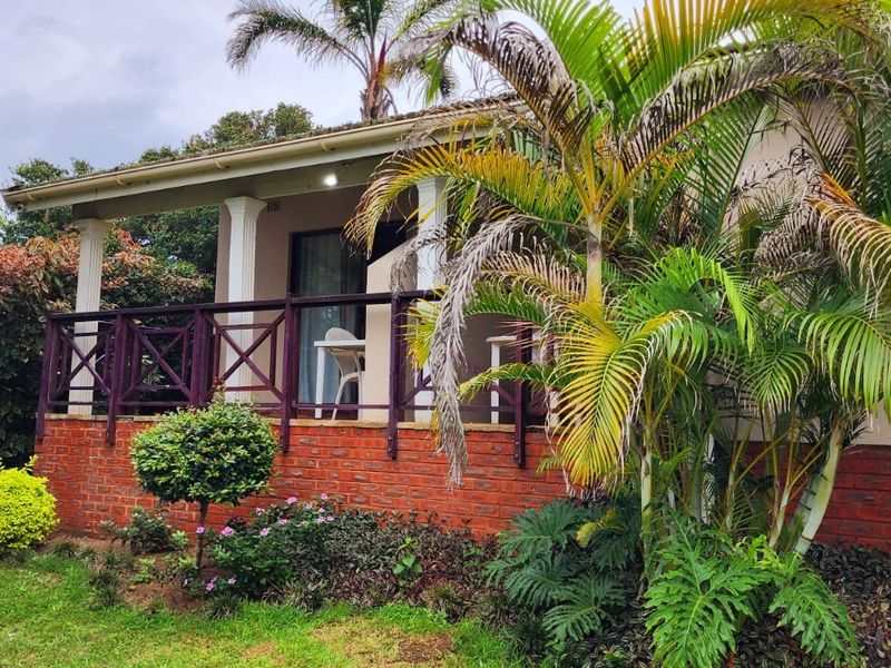 A covered patio area featuring a brick base, wooden balustrade, and support columns, set within a landscaped garden environment.