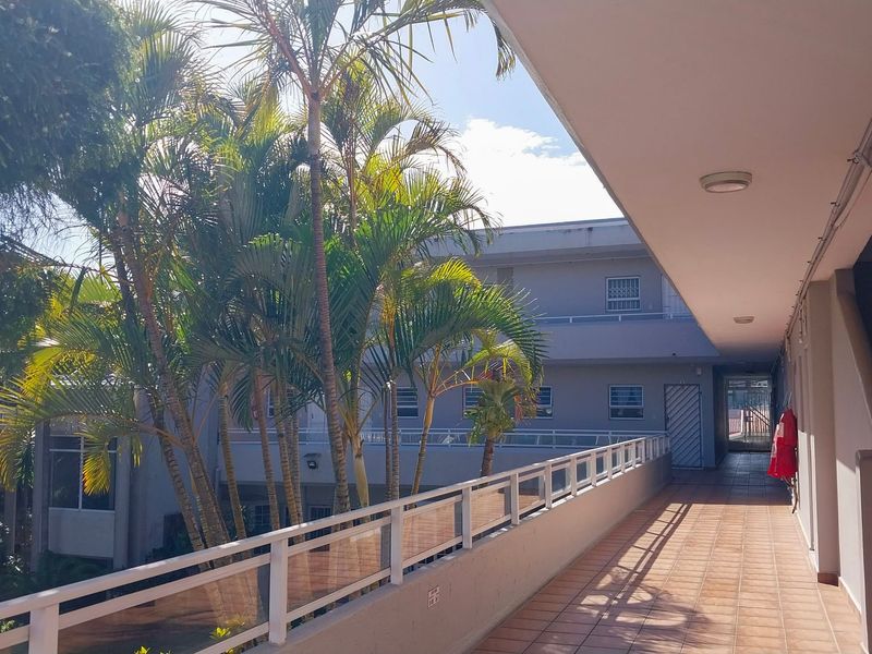 An open-air apartment walkway featuring tiled flooring, a metal balustrade, and views of the surrounding complex greenery.