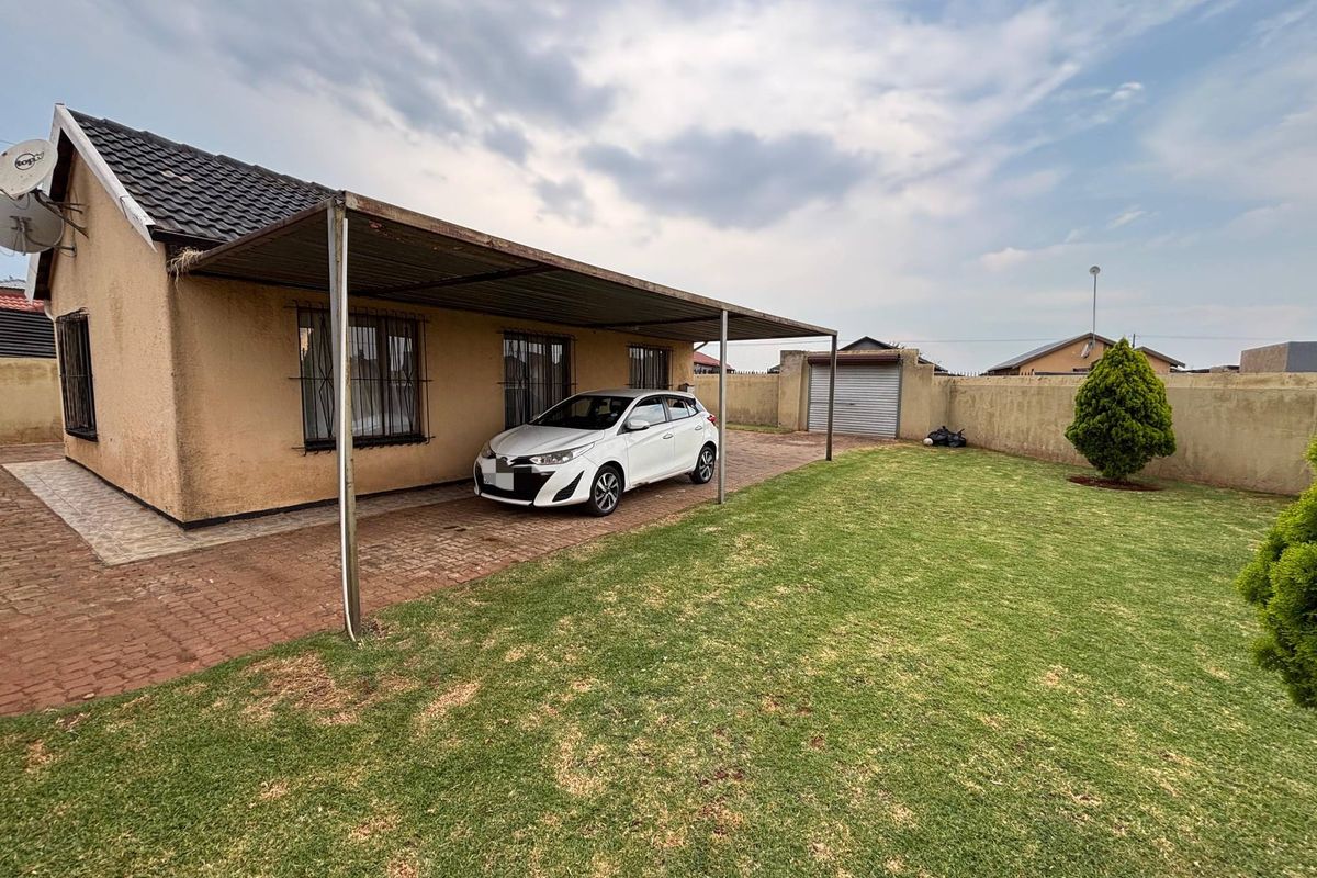 Rear exterior view of the property featuring a paved driveway, metal carport, and a spacious garden area enclosed by a perimeter wall.