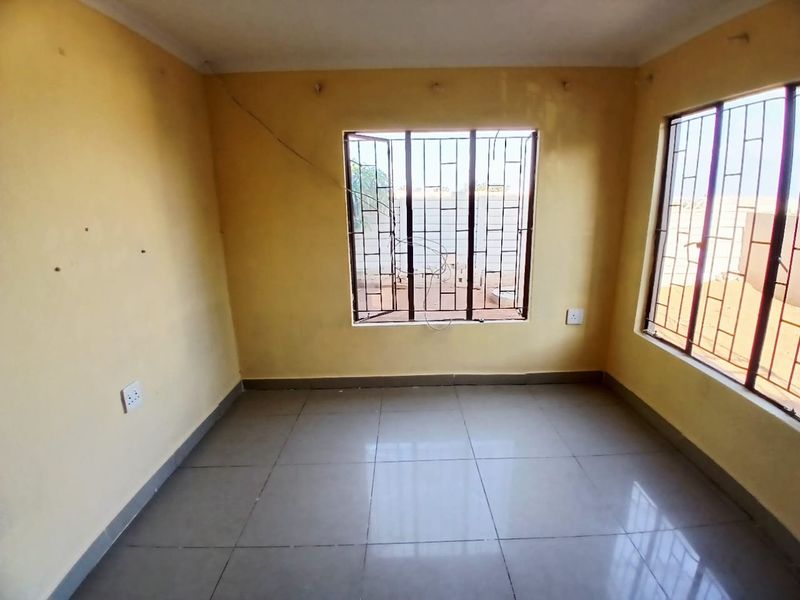An empty bedroom featuring tiled flooring, security bars on the windows, and yellow painted walls with visible surface cracks.