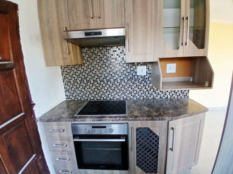 A compact kitchen featuring wood-look cabinetry, a mosaic tile backsplash, integrated electric hob, oven, and extractor fan.