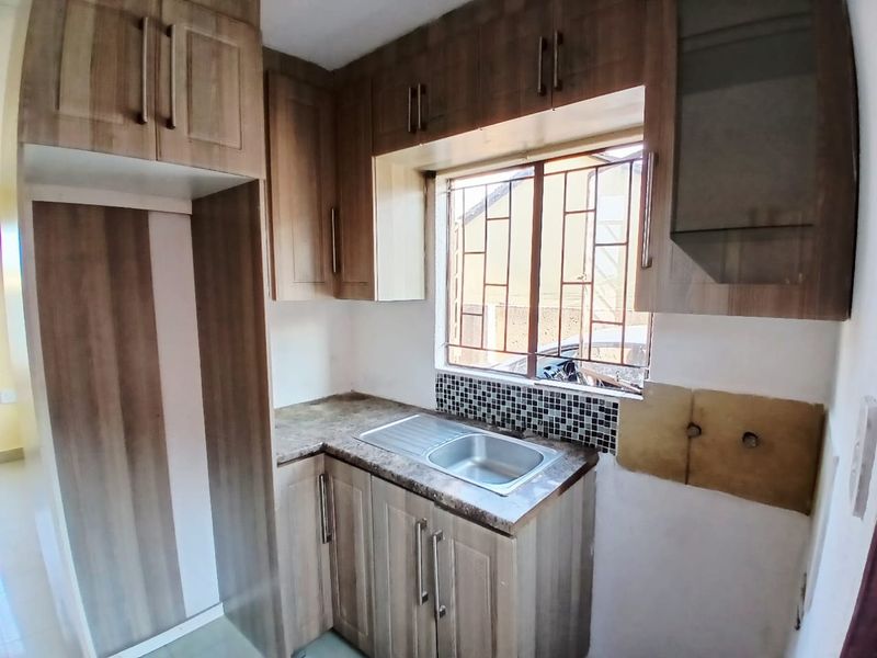 Compact kitchen featuring wood-look laminate cabinetry, a stainless steel sink, and mosaic tile backsplash, with an unfinished wall section requiring attention.