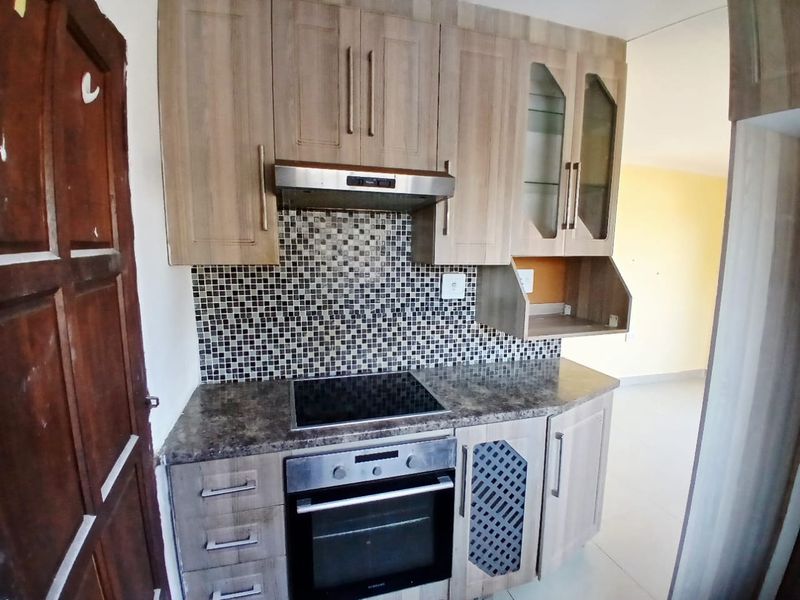 A compact kitchen featuring wood-look laminate cabinetry, a mosaic tile backsplash, built-in oven, electric hob, and an integrated extractor fan.