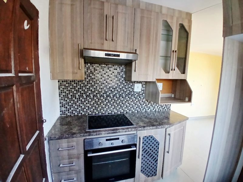 Compact kitchen featuring wood-look cabinetry, mosaic tile backsplash, integrated electric hob, oven, and extractor fan, offering a functional layout for meal preparation.