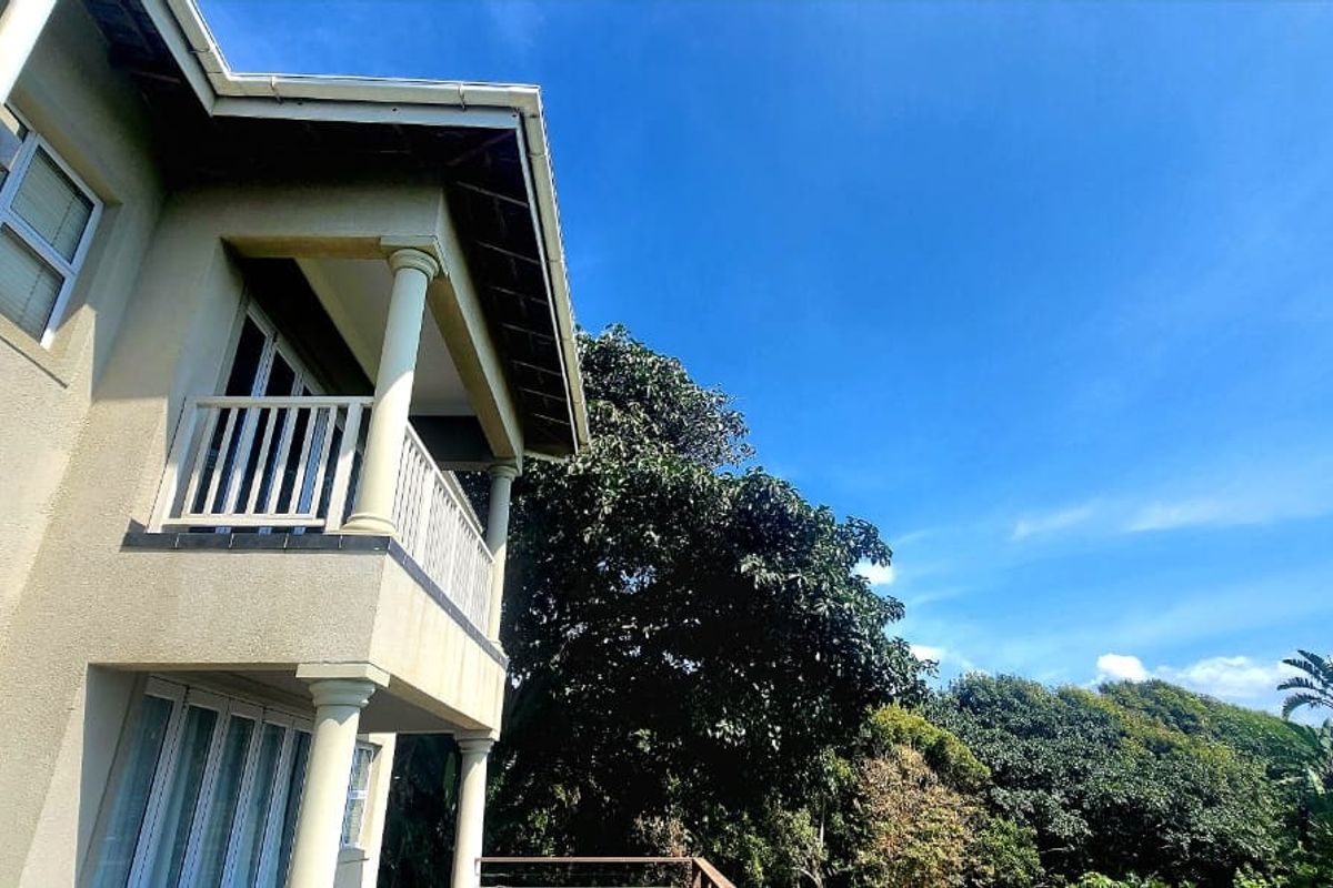 A view of the rear exterior facade featuring a balcony with columns and railings, overlooking lush greenery under a clear blue sky.