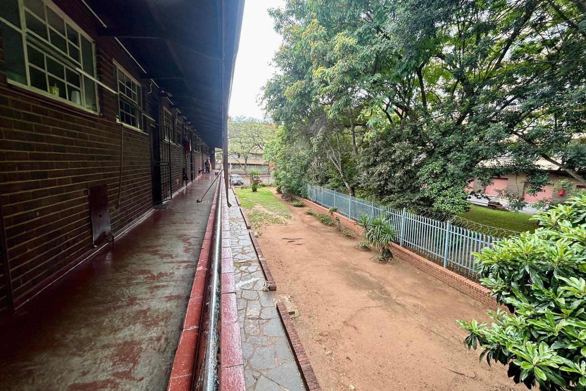 An exterior balcony walkway overlooking a communal garden area, featuring brick wall construction and metal security fencing.
