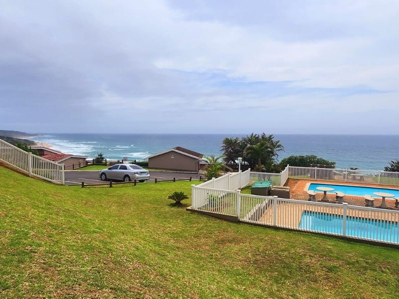 Rear exterior view of the property featuring a swimming pool, paved deck, white fencing, and expansive lawn with ocean views.