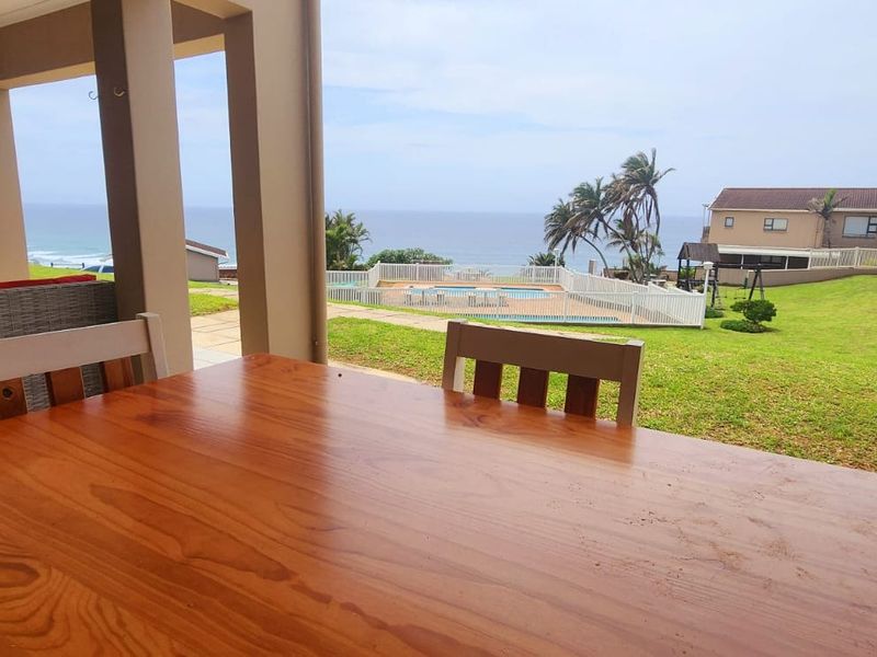 A covered patio area featuring tiled flooring and concrete pillars, offering scenic views of the communal garden, swimming pool, and the ocean beyond.