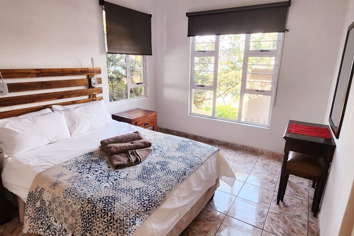 A well-lit bedroom featuring tiled flooring, large aluminium-framed windows with roller blinds, and a neutral interior finish.